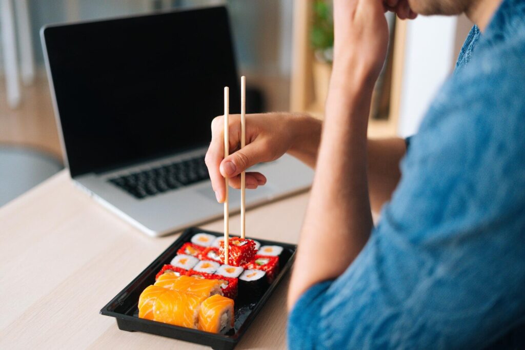 Closeup back view unrecognizable young man eating sushi rolls with chopsticks sitting working desk with laptop home office 482921 7403 Closeup back view unrecognizable young man eating sushi rolls with chopsticks sitting working desk with laptop home office 482921 7403